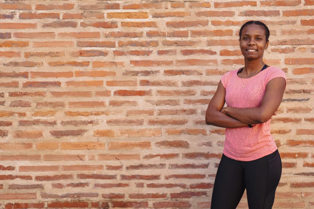 Coach sportive posant avec assurance devant un mur en briques toulousain, vêtue d’un t-shirt rose et d’un legging noir – coaching à domicile Toulouse.