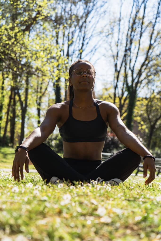 Femme assise en tailleur dans l’herbe, en posture de méditation, les yeux fermés – séance de coaching bien-être ou hypnose en extérieur à Toulouse.