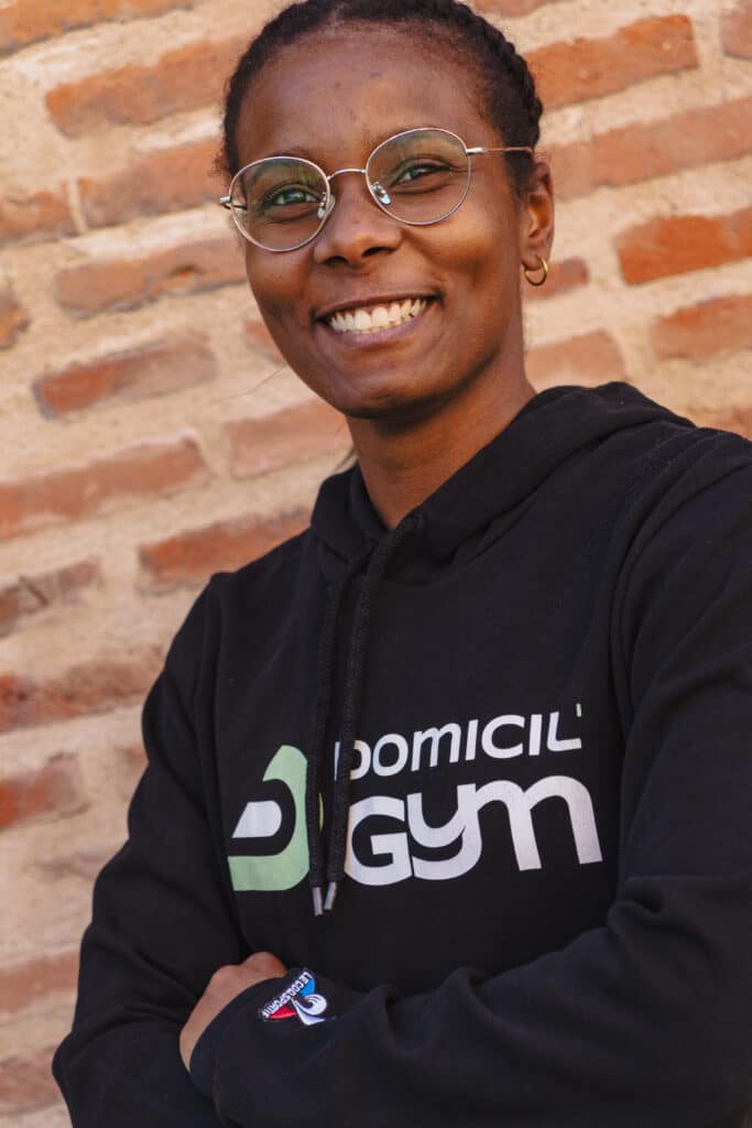 Coach sportive souriante portant un sweat à capuche Domicil’Gym, posant devant un mur en briques – coaching à domicile personnalisé.