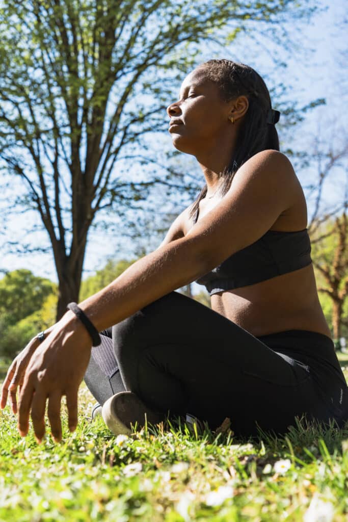 Femme en posture de méditation assise dans l’herbe, les yeux fermés et le visage tourné vers le soleil – séance de recentrage et relaxation en pleine nature.