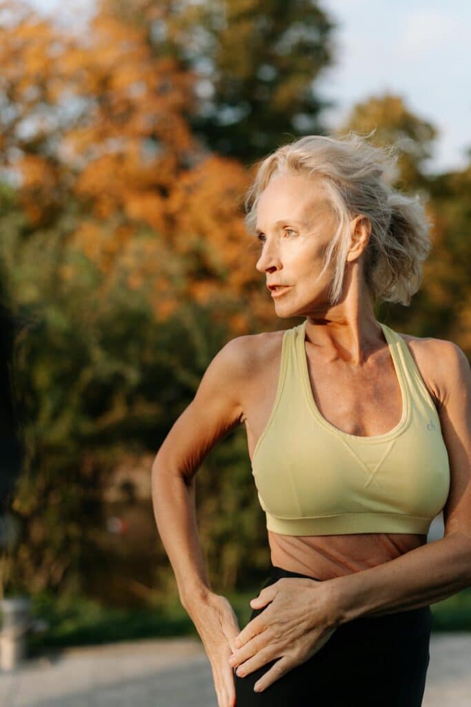 Femme senior athlétique aux cheveux gris, regardant sur le côté, portant une brassière de sport jaune. Elle est à l'extérieur, sous le soleil d'automne.
