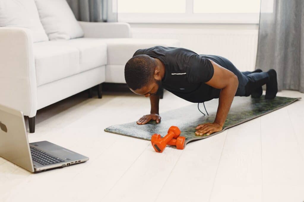 Homme noir faisant un push-up sur un tapis de yoga, devant un laptop ouvert et des haltères orange, dans un salon moderne.